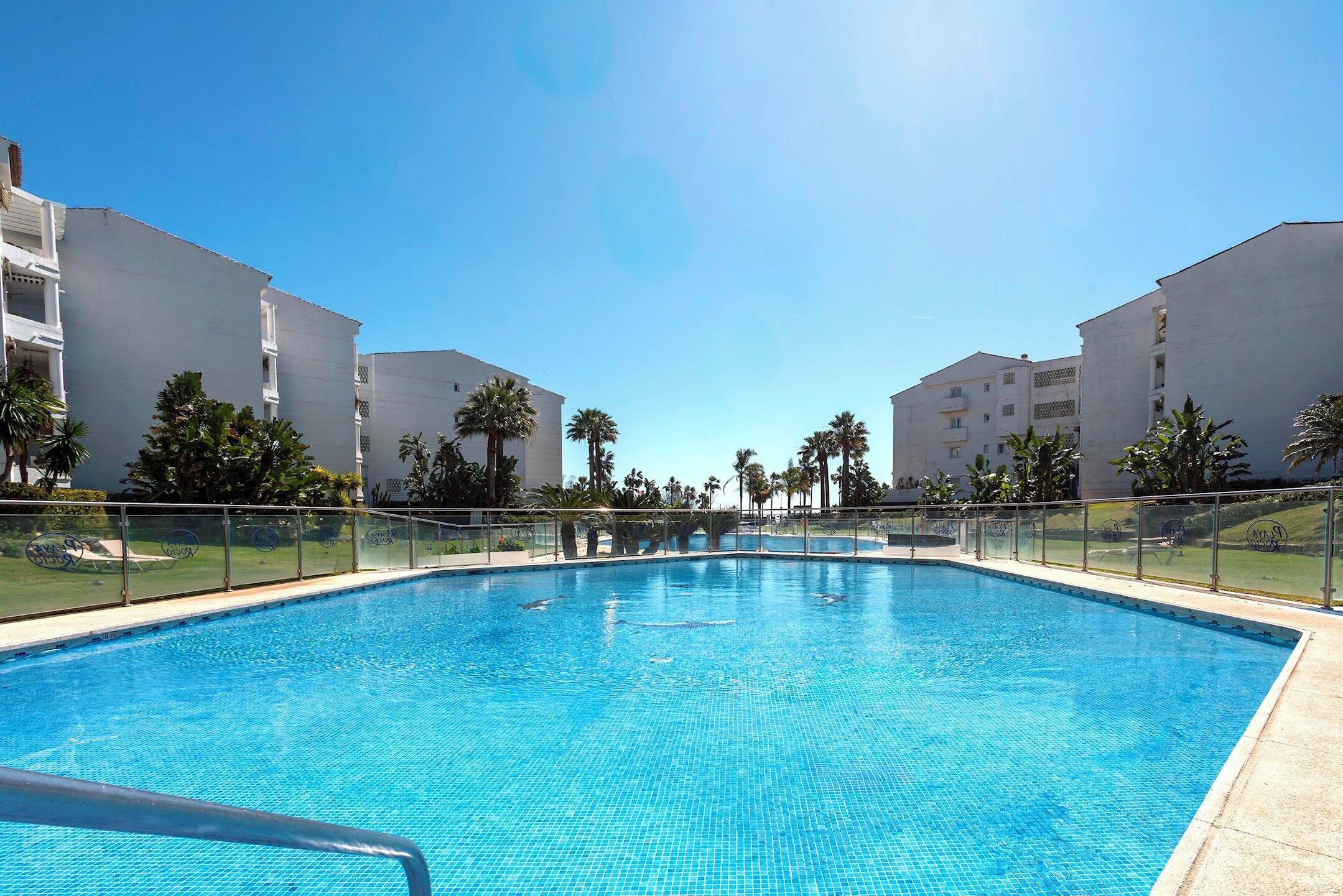 A swimming pool with a clear blue water, surrounded by white buildings and palm trees.