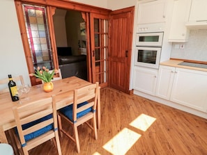 A wooden dining table with chairs, a kitchen with white cabinets and double ovens, and a glass door entrance.