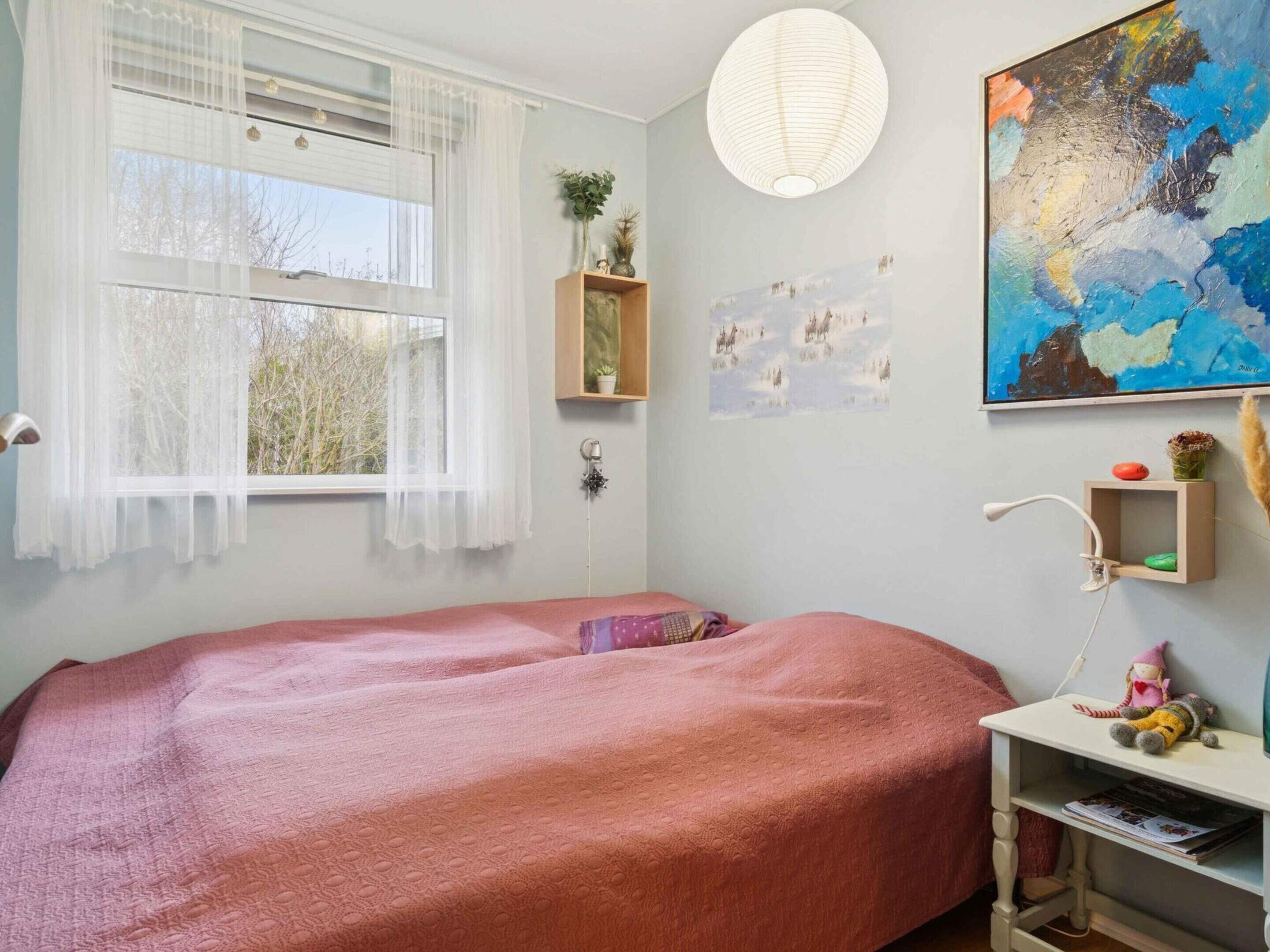 Une chambre à coucher avec un lit, une fenêtre ornée de rideaux légers, une lampe fixée au mur, une étagère avec des objets décoratifs et un tableau.