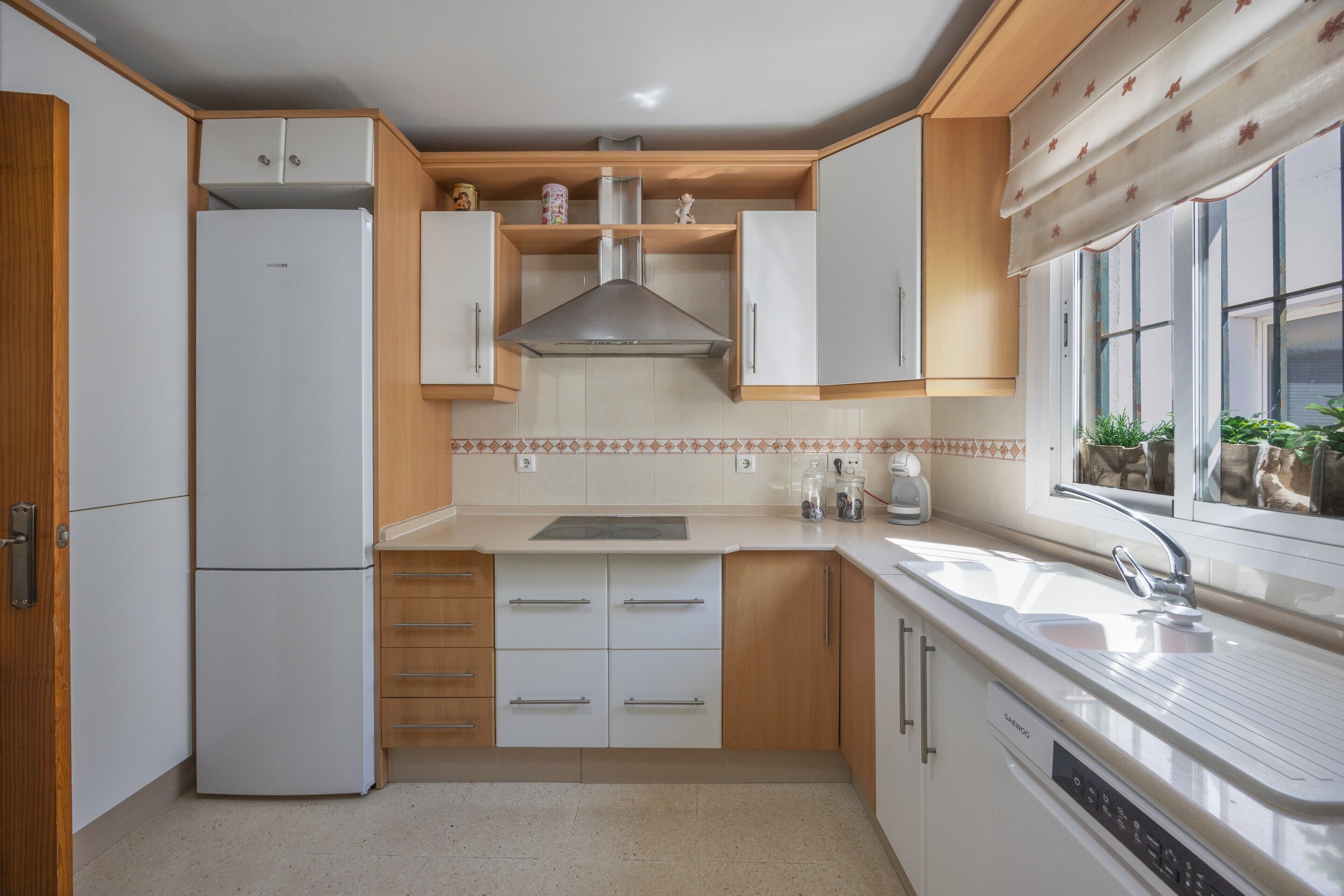 A compact kitchen with white cabinets, a stainless steel range hood, a built-in dishwasher, and a window with a view of greenery.