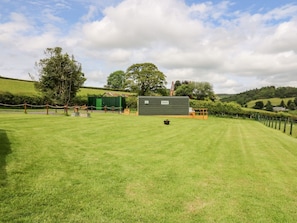 A grassy field with a green building, a fenced area, and trees in the background. - Hwyl, Llandrindod Wells (Llandrindod Wells)