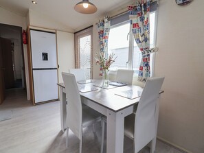 A compact kitchen with a refrigerator, a dining table with chairs, and a window with patterned curtains. - Caravan Cp10 (Prestatyn)