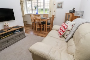 A living room with a beige sofa, wooden dining table, chairs, a TV, a bookshelf, and a window with curtains.