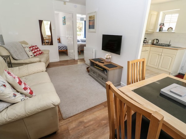 A living room with a sofa, a TV, a dining table with chairs, a kitchen area with cabinets, and a doorway leading to another room.