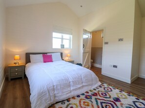A bedroom with a bed, two bedside tables with lamps, a window, a door leading to a bathroom, and a patterned rug. - The Stables, Tafarn-Y-Gelyn (Mold)