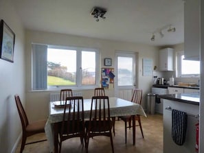 A dining area with a table and chairs, a kitchen with cabinets and appliances, and a view through the windows. - Charming Godolphin Retreat in Polzeath (Wadebridge)