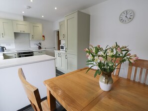 A kitchen with a wooden dining table, a vase of flowers, and a clock on the wall. - Cottage on the Green, Gwynedd. (Caernarfon)