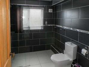 A bathroom with dark tiles, a white toilet, a window with blinds, and a towel rack.
