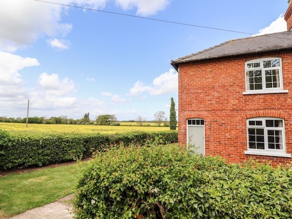A red brick house with a white door and window, surrounded by well-maintained hedges and a grassy field.
