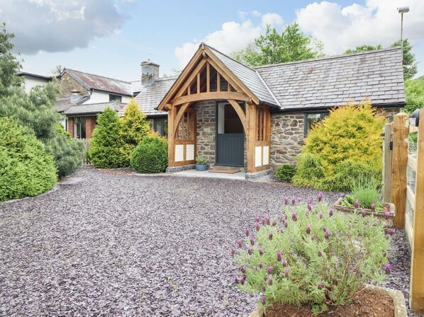 A stone building with a wooden porch, gravel driveway, and surrounding greenery.