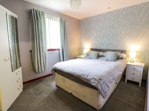 A bedroom with a bed, two bedside tables, a nightstand, a window with curtains, and a wall with a tree pattern. - Torkeld Holiday Cottage, Garlieston (Newton Stewart)