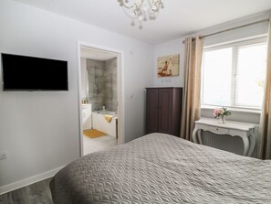 Un dormitorio con cama, televisor montado en la pared, armario, ventana con cortinas y baño a la vista a través de una puerta abierta.