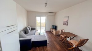 Ein modernes Wohnzimmer mit einer Couch, einem Fernseher, einem Holzesstisch und einem Balkon mit Gartenmöbeln.