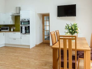 A modern kitchen with white cabinets, a microwave, and an oven. There is a dining table with chairs and a potted plant. A television is mounted on the wall.