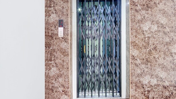 Une porte munie d’une grille de sécurité en métal, un mur à la texture évoquant le marbre, et un panneau de commande avec un bouton et un écran.
