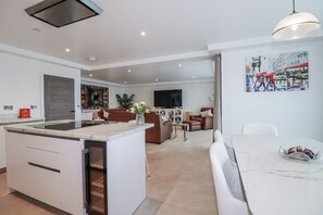 A modern living room with a sofa, a dining table with fruit, a kitchen island, and a TV. - 13 Cliff Edge, Newquay (Newquay)