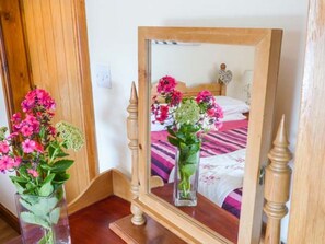 A wooden-framed mirror reflecting a bed with a pink and white striped bedspread, a heart-shaped decoration on the wall, and two vases with flowers.
