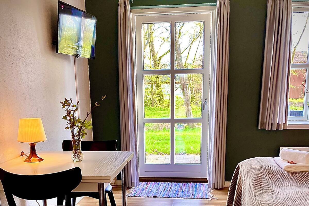 A hotel room with a dining table, chairs, a TV, a bed with a striped bedspread, and a view of the outdoors through a glass door.