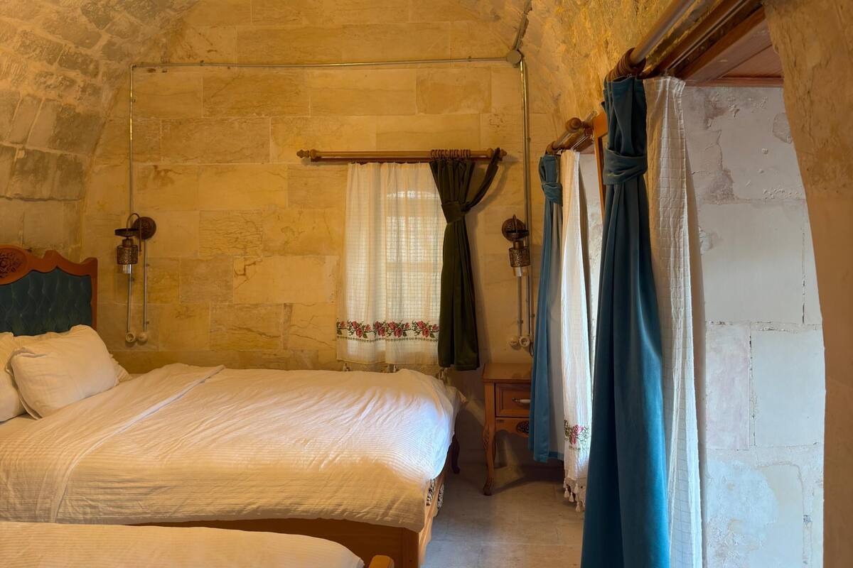 Una habitación con dos camas, paredes de piedra, cabecero de madera, una ventana con cortinas y una lámpara colgante.