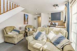 A living room with two armchairs, a sofa, a staircase, a kitchen area, and a clock on the wall.