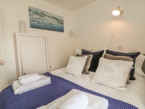 Un lit bien fait, avec une literie blanche et bleue, un tableau représentant un paysage côtier et un luminaire fixé au mur.