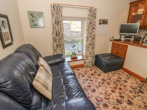 A room with a black leather sofa, a small wooden cabinet, a television, and a window with floral curtains. - Sycamore Cottage, Hawes (Sedbergh)