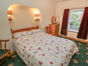 A bedroom with a floral bedspread, a wooden dresser, a chair, and a window with curtains. - Sycamore Cottage, Hawes (Sedbergh)