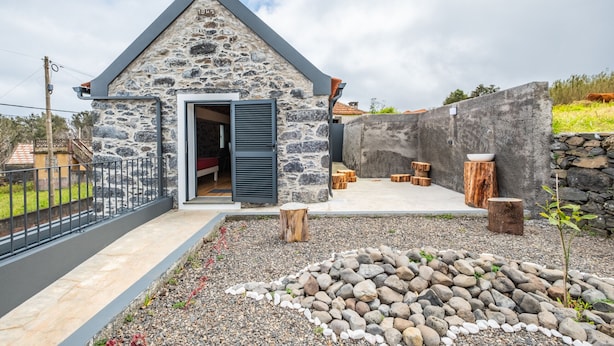 A stone building with a sloped roof, a door, and a small outdoor seating area with wooden stools. - N T House by Homie in Porto Moniz (Porto Moniz)