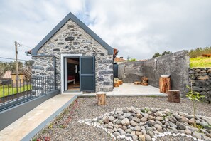 A stone building with a sloped roof, a door, and a small outdoor seating area with wooden stools. - N T House by Homie in Porto Moniz (Porto Moniz)