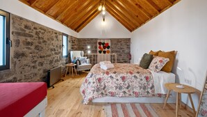 A bedroom with a sloped wooden ceiling, a stone wall, a bed with a floral bedspread, a chair, and a small table. - N T House by Homie in Porto Moniz (Porto Moniz)