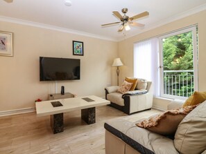 Una sala de estar con una televisión de pantalla plana, ventilador de techo, sofá, mesa de centro y un ventanal con vistas a zonas verdes.