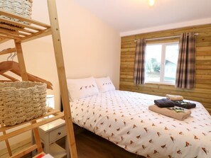 Un dormitorio con una litera de madera, una cama individual con ropa de cama estampada, una ventana con cortinas y una mesita de noche pequeña.