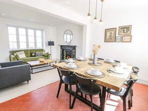 Een eetgedeelte met een houten tafel voor zes personen, omringd door stoelen, een open haard en een bank op de achtergrond.