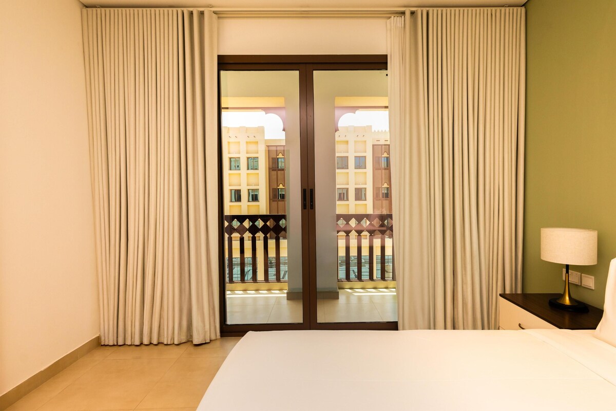 A hotel room with a bed, a nightstand with a lamp, and a view of a building through a glass door.
