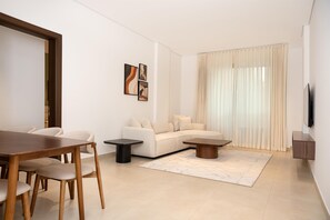 A modern living room with a sofa, a dining table with chairs, a coffee table, and a flat-screen TV mounted on the wall. - Tulip Residences Muscat The Village (Seeb)