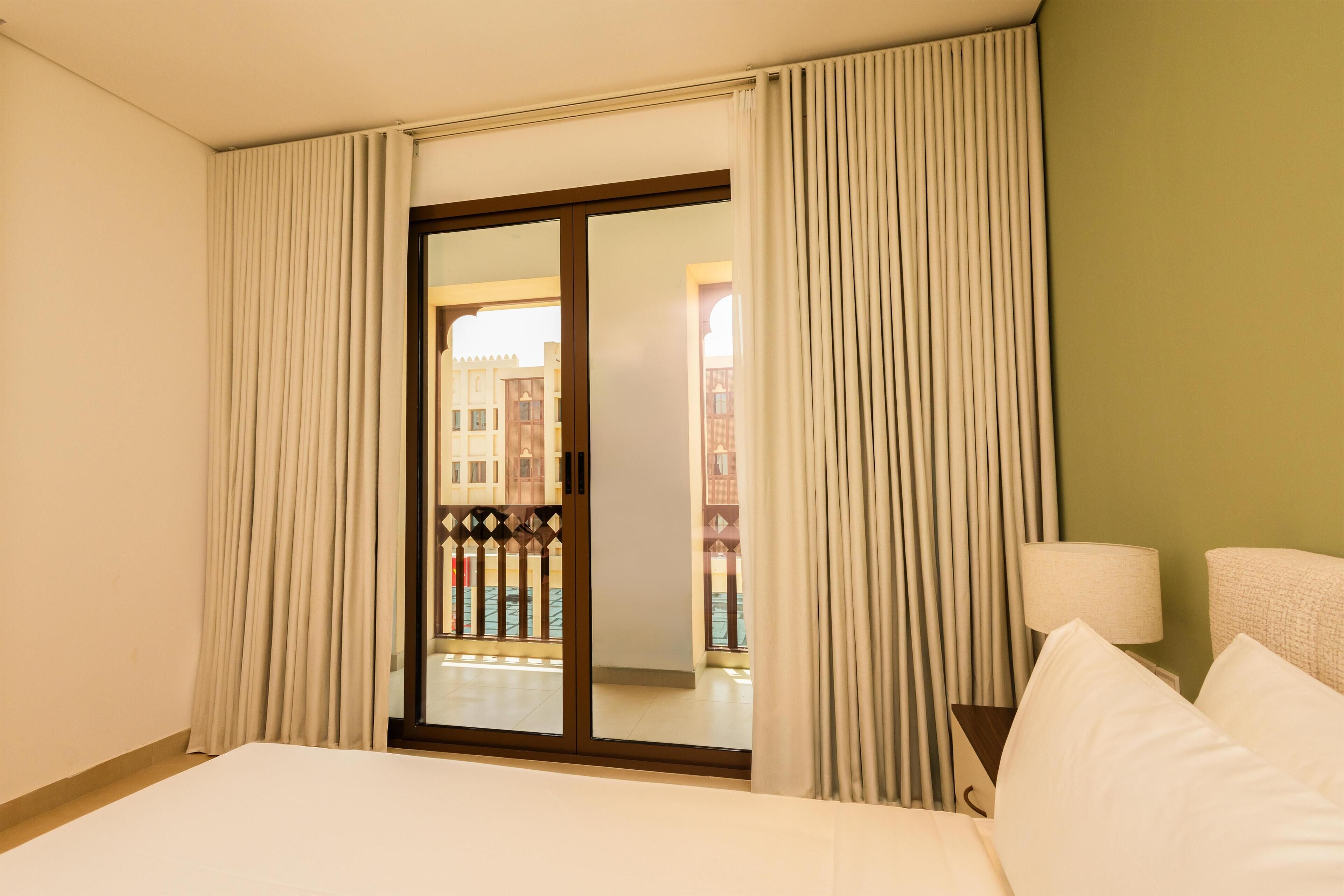 Quarto de hotel com cama, mesa de cabeceira, abajur e vista para um edifício através de uma porta deslizante.