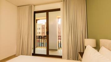 Quarto de hotel com cama, mesa de cabeceira, abajur e vista para um edifício através de uma porta deslizante.