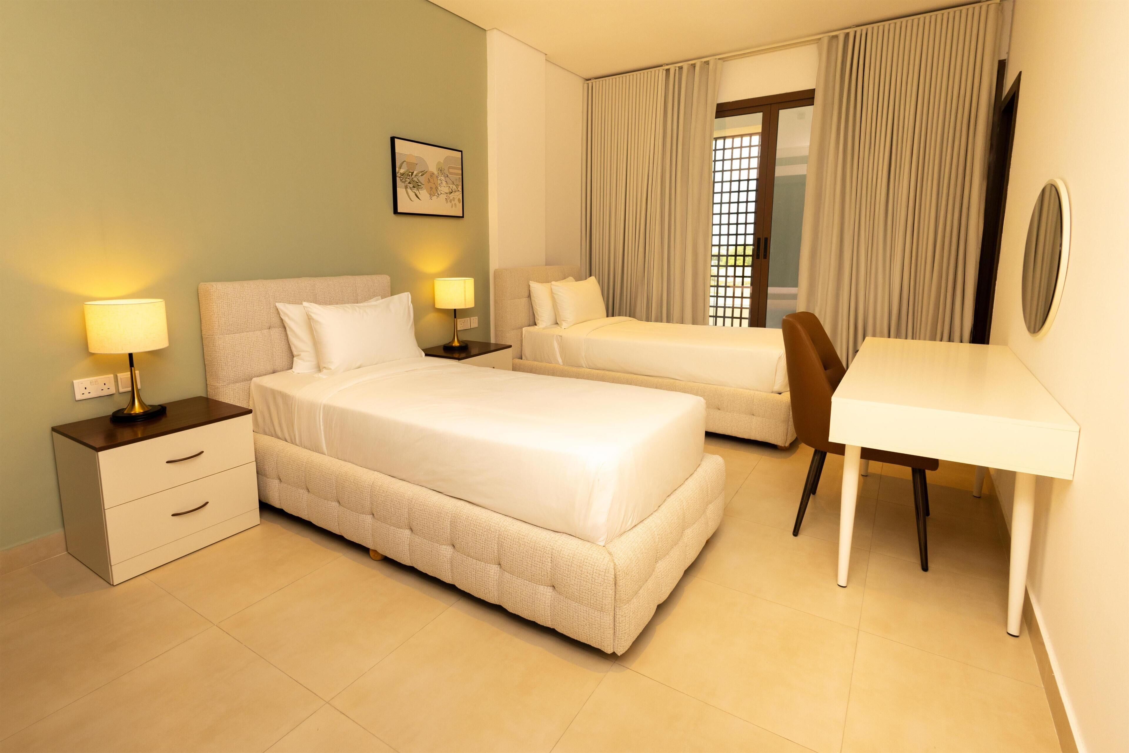 A hotel room with two beds, a desk, and a chair. There is a window with curtains, a bedside table with a lamp, and a wall-mounted picture.