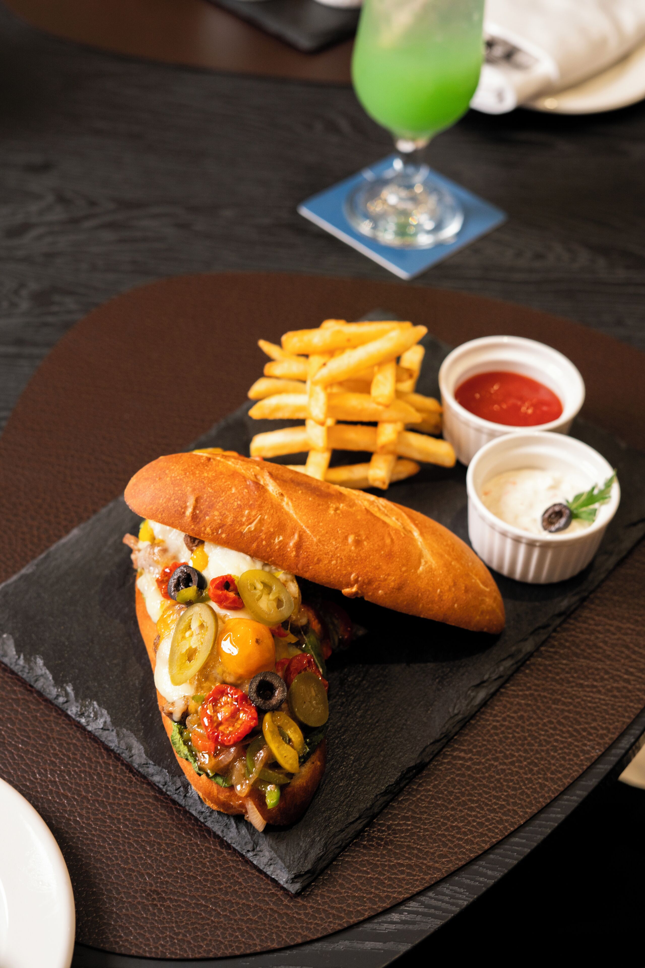 Un sándwich con diversos ingredientes, una porción de papas fritas y dos recipientes pequeños de salsa sobre un plato de pizarra.