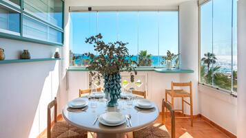 Una mesa de comedor redonda con sillas, un jarrón con flores y vistas al océano a través de amplios ventanales.