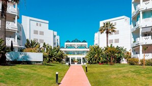 A pathway leading to a modern building with a central glass facade, surrounded by lush greenery and palm trees. - Beachfront Apartment in Puerto Banus Playa Rocio (Marbella)