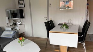 Ein modernes Wohnzimmer mit einer Couch, einem Couchtisch mit Blumen, einem Fernseher, einem Esstisch mit Stühlen und einer Tür.