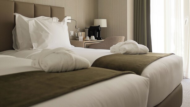 A neatly made bed with white linens and brown accents, a bedside table with a lamp, a coffee maker, and a bottle of water. - OMID Boutique (Lisbon)