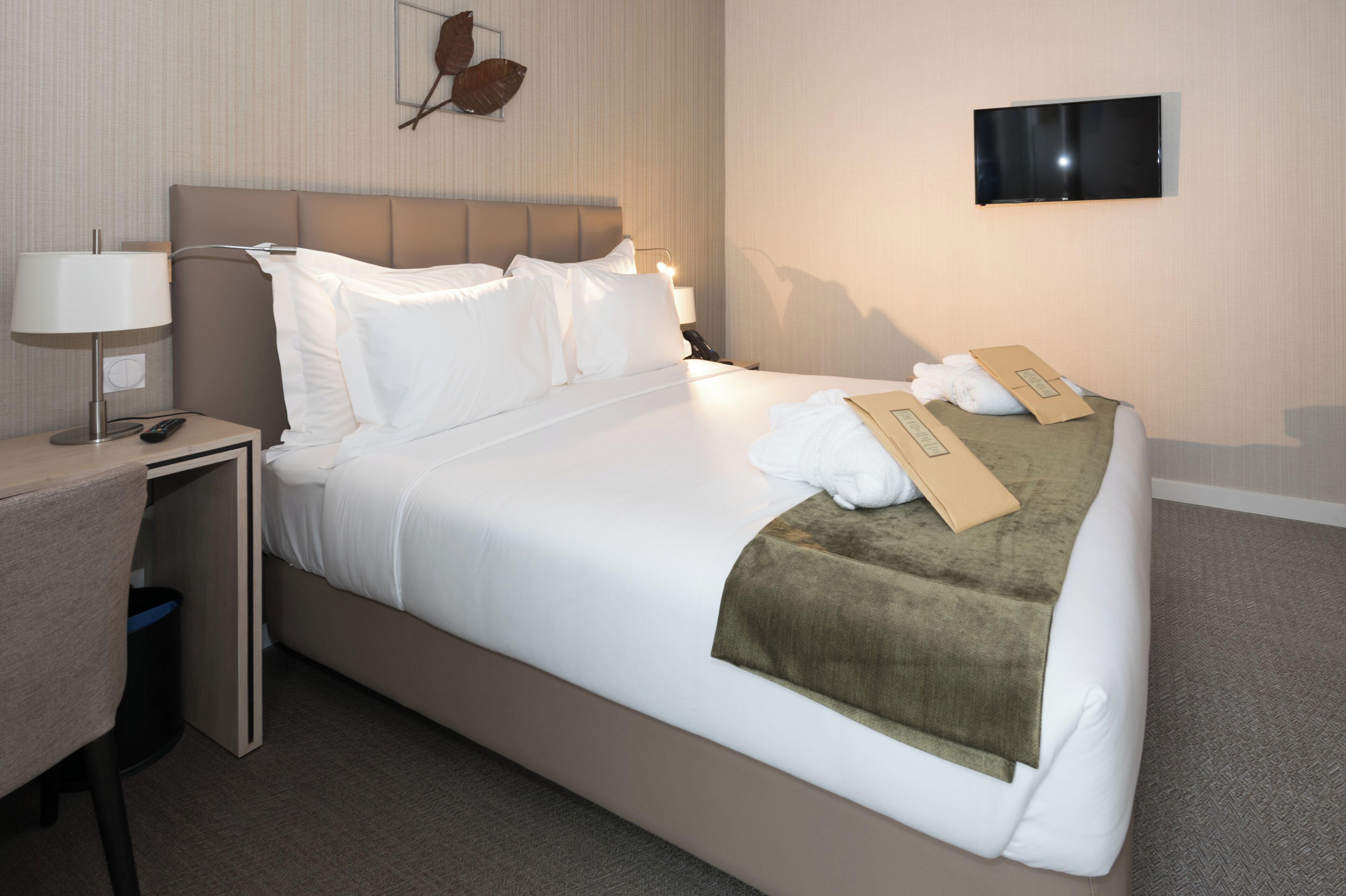 A hotel room with a bed, a desk, a lamp, a trash bin, and a flat-screen TV on the wall.