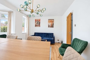 Ein modernes Wohnzimmer mit einem Holzes Esstisch, einem blauen Sofa und zwei grünen Sesseln. An der Wand befinden sich zwei Retro-Partyplakate.