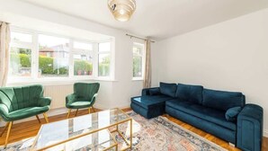 Un salon avec un canapé bleu, deux fauteuils verts, une table basse en verre et un tapis à motifs.