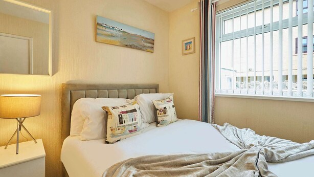 Ein Hotelzimmer mit Bett, Nachttisch, Lampe, einem Fenster mit Gitter und einem Bild an der Wand.