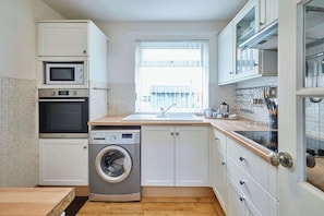 A compact kitchen with a washing machine, a microwave, and a sink with a window above. - Host & Stay | The Garden Flat, Seahouses (Seahouses)