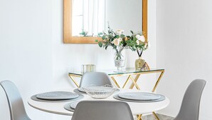 Een eethoek met een ronde tafel, grijze stoelen, een glazen vaas met bloemen en een spiegel met gouden lijst aan de muur.