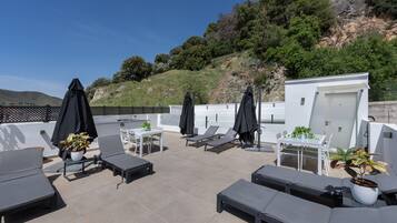 Une terrasse sur le toit, aménagée avec des chaises longues, des tables et des parasols, offrant une vue sur une colline.